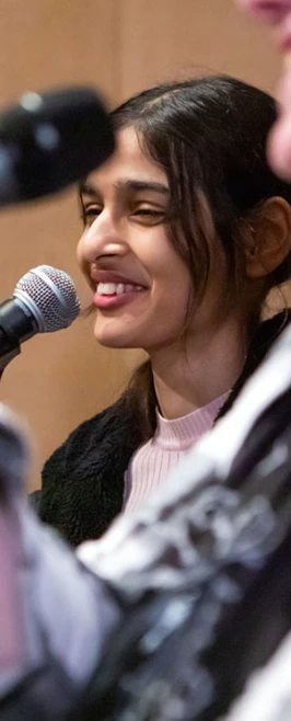 A student singer in a pink sweater smiling while singing into a microphone during a band practice session. A student singer in a pink sweater smiling while singing into a microphone during a band practice session.